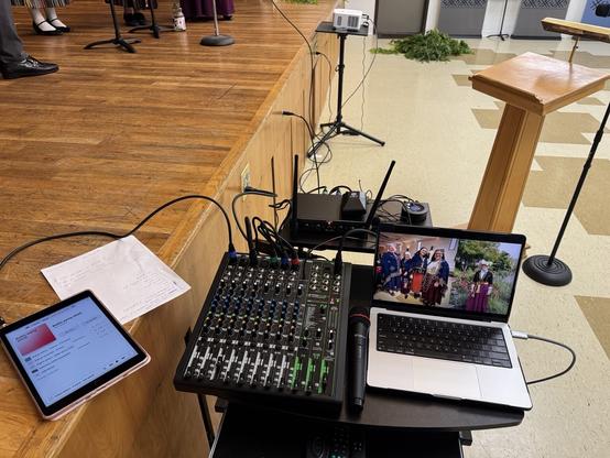 In front of a stage with a small rolling card with my audio/video battle station with a 16 track mixer, a four-mic wireless systems, a laptop, and a tablet. In back is a projector, some feet and microphone based, a lectern, anf and a big pile of branches for decorations.