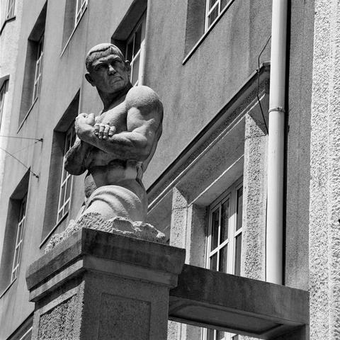 The black and white image shows a muscular stone statue of a man standing on a pedestal in front of a building. The figure has a stern expression and is posed with arms crossed over his chest, highlighting his defined physique. The background features tall windows and textured walls, suggesting an institutional or historical setting.