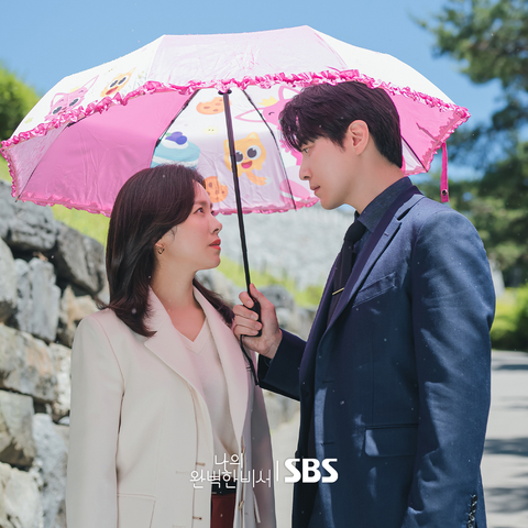 Han Ji Min in a white suit looking up at Lee Jun Hyuk in a dark grey suit holding a pink, frilly umbrella with cartoon characters on it, over both of them from the 2025 South Korean drama "Love Scout."