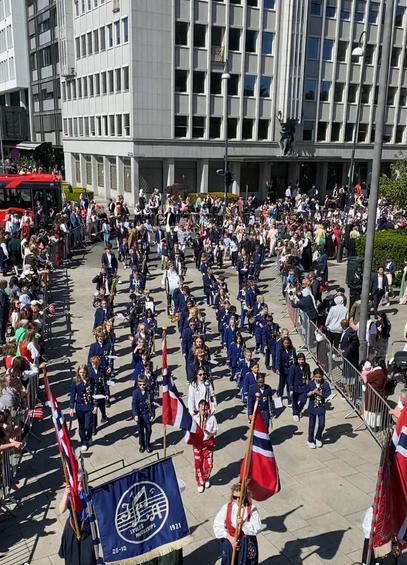 Korps under marsjering sett oppefra, de har blå uniformer og er stilt opp i rekker på 5. De går mellom sperregjerder med folkemengde på begge sider.