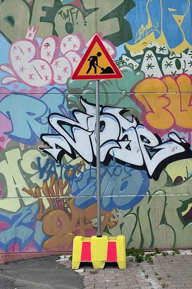 A "roadworks" sign in front of a wall covered with street art tags. The framing lets the viewer be tricked by the fact that the sign may refer to the art.