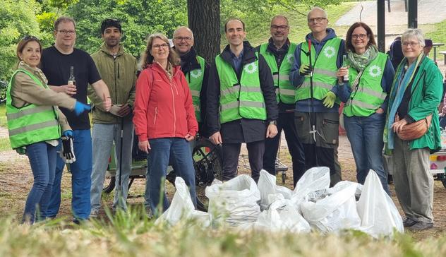 Zehn Personen, sechs mit grüner Weste, stehen auf einer Wiese, davor einige weiße Müllsäcke.
