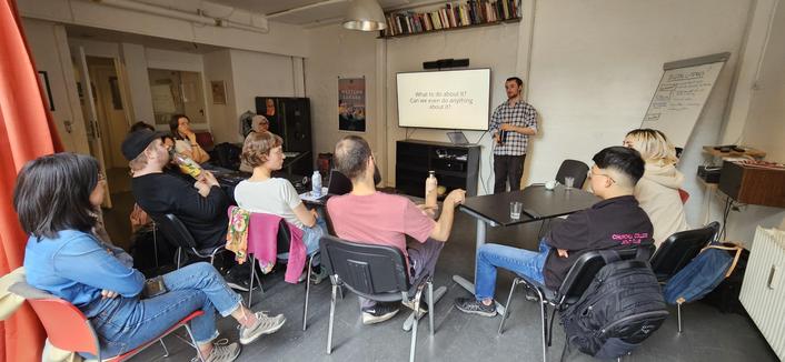 A room full with 10 people in a semi circle around one person presenting at a screen