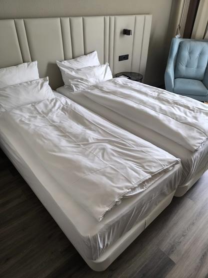 Das Bild zeigt ein geräumiges Hotelzimmer mit zwei Einzelbetten, die nebeneinander stehen. Die Betten sind mit weißen Bettwäsche und Kissen bedeckt, die sauber und ordentlich aussehen. Die Kopfstütze der Betten ist in einem hellen Beige gehalten und hat eine gepolsterte, vertikale Struktur. An der Wand neben den Betten befindet sich ein schmaler Nachttisch mit einer Lampe und einer Steckdose. Rechts im Bild ist ein hellblauer Sessel mit gepolsterten Armlehnen zu sehen. Der Boden besteht aus dunklem Holzverkleidung, was dem Raum eine warme Atmosphäre verleiht. Die Fenster sind mit hellen Vorhängen ausgestattet, die das natürliche Licht hereinlassen.