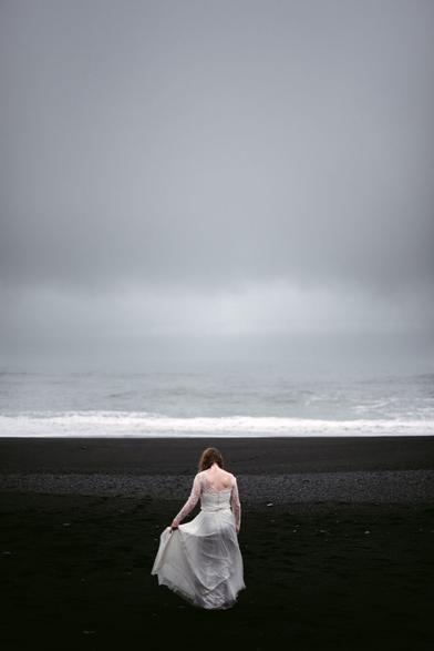 This atmospheric photograph by Elopement Photographer Sturmsucht captures a solitary bride walking across Iceland's distinctive black sand beach. She is seen from behind, wearing a flowing white lace wedding gown with delicate long sleeves and an open back design. The bride is gently holding the edge of her dress, which appears to be moving slightly in the breeze.  The landscape creates a dramatic contrast between her white dress and the deep black volcanic sand beneath her feet. Above, heavy gray skies dominate the scene, creating a moody, overcast atmosphere. In the distance, gentle waves of the ocean can be seen breaking on the shore with white foam against the dark beach.  The overall mood is contemplative and serene, capturing a quiet moment during what appears to be an elopement ceremony. The minimalist composition emphasizes the solitude of the bride against the vast, stark Icelandic landscape, creating a poetic and emotionally evocative image.
