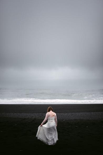 This atmospheric photograph by Elopement Photographer Sturmsucht captures a solitary bride walking across Iceland's distinctive black sand beach. She is seen from behind, wearing a flowing white lace wedding gown with delicate long sleeves and an open back design. The bride is gently holding the edge of her dress, which appears to be moving slightly in the breeze.  The landscape creates a dramatic contrast between her white dress and the deep black volcanic sand beneath her feet. Above, heavy gray skies dominate the scene, creating a moody, overcast atmosphere. In the distance, gentle waves of the ocean can be seen breaking on the shore with white foam against the dark beach.  The overall mood is contemplative and serene, capturing a quiet moment during what appears to be an elopement ceremony. The minimalist composition emphasizes the solitude of the bride against the vast, stark Icelandic landscape, creating a poetic and emotionally evocative image.