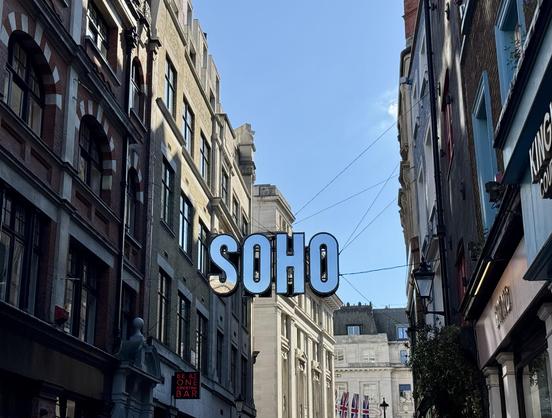 Angled image of building facades in London’s Soho, centered on a large sign reading SOHO hanging across a street