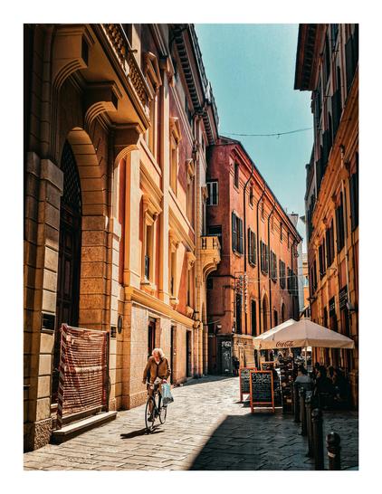 The photo is of a narrow, sunlit street in Bologna. Tall, ochre/yellow-coloured buildings are on both sides of the street. They are highlighted from the strong sunlight. There is an elderly man with white hair is riding a bicycle down the street, carrying a shopping bag in the front left of the foreground. In the background, a cafe with a Coca-Cola branded umbrella is visible, with people seated at tables.