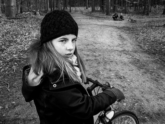 Mädchen auf dem Fahrrad, das in die Kamera schaut. Im Hintergrund verprügeln ein paar Jungs ein auf dem Boden liegendes Kind.