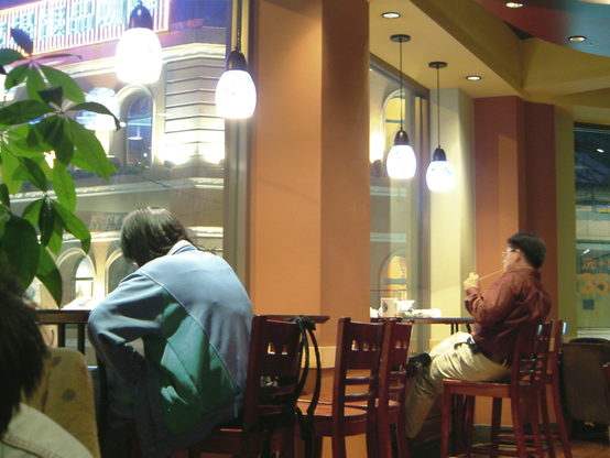 Inside this cafe, people sit at tables near the large glass front facing the busy street. Brightly lit European style facades are visible, as are Chinese chracters in various places (window decor, advertising). Various drinks and other items are on the tables, and everyone seems to busy themselves with reading or writing. There are no smartphones or laptops visible.