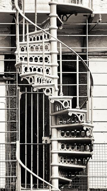 A black and white photograph shows a vintage white metal spiral staircase attached to a building with wooden plank siding. The staircase curves upwards with ornate circular and geometric cutouts in the steps and railings. A dark, barred door is visible behind the lower section of the staircase.