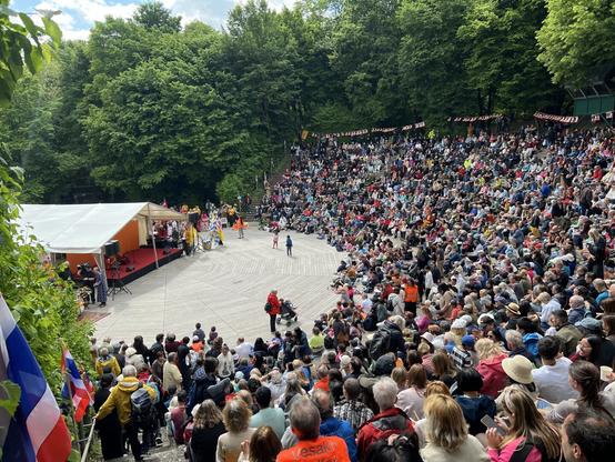 Eine gefüllte Tribüne im Oval der Seebühne im Westpark München mit grün blühenden Bäumen im Hintergrund und derzeit noch einer leeren Bühne mit weißem Vorzelt während der Vorbereitung.