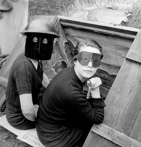 SW Foto
2 Frauen sitzen auf Zeitungspapier. Sie schauen die Fotografin an. Sie tragen schwarze Masken, die surreal aussehen. eine trägt einen Helm.