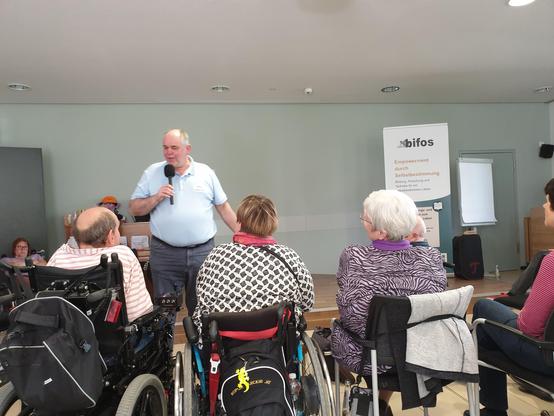 Ottmar mile paul spricht ins Mikrofon,  menschen hören zu. Einige sitzen im Rollstuhl