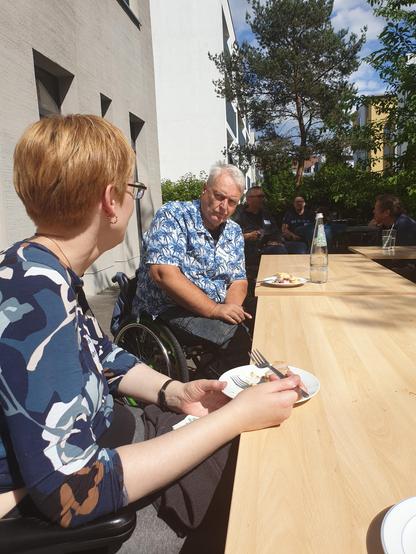 Zwei Rollstuhlnutzende an einem Tisch draußen in der Sonne