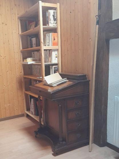 Le davenport vu de trois quart à côté d'une bibliothèque rudimentaire en pin massif, remplie de livres de poches. Les deux meubles sont dans l'angle d'une pièce aux murs recouverts de lambris