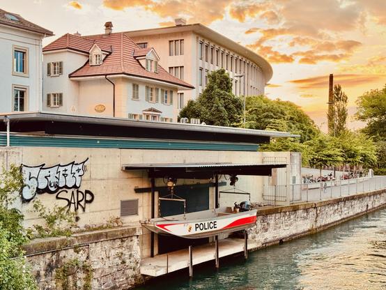 In the Swiss town of Thun, a small police boat is suspended above the River Aare (ARR) in a sheltered boat lift structure attached to the riverbank. The boat, marked “POLICE,” hangs neatly above the water, ready for deployment. The concrete wall of the structure is covered in graffiti. Nearby, there's a charming old house with a red-tiled roof, and behind it stands a large modern building partially covered by lush green trees. A row of bicycles lines a path along the water. The sky glows with a warm, golden sunset, casting a peaceful yet slightly surreal atmosphere over the riverside scene.