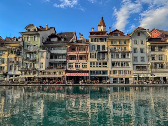 A row of colorful, tightly packed historic buildings lines the river Aare in Thun, Switzerland. The facades range from pale yellow and mint green to pastel blue and red, with charming balconies and shuttered windows. At the base, cafés and restaurants host people dining outdoors by the water. The river reflects the buildings perfectly in its calm, emerald green surface. Above, the sky is bright blue with scattered clouds.