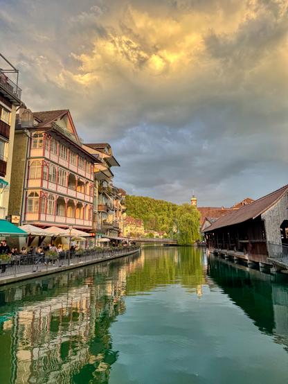 This view captures the same river further along, just before sunset. On the left, more elegant wooden houses and a busy café terrace hug the riverbank. On the right is a traditional covered wooden bridge with a sloped roof. The sky is dramatic with golden clouds breaking through, casting a warm glow over the green hills and clocktower in the background. The water reflects everything in soft, rippling detail.