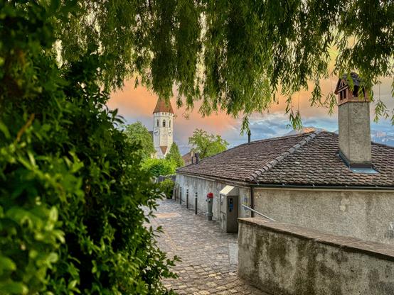 A cobbled alley gently slopes down between old, tiled rooftops in the town of Thun. Framed by leafy branches, the scene feels hidden and peaceful. In the background stands a medieval white church tower with a red pointed roof. Just beyond the rooftops, the distant Alps rise quietly under a sky glowing with sunset colors. The atmosphere is serene and timeless.