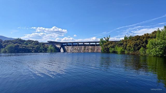 Fotografía en la que se ve el muro que separa (y comunica) el Embalse de Los Arroyos con el Embalse de Valmayor. 
A ambos lados del muro se puede ver mucha vegetación de árboles, de muchos de ellos solo se aprecian sus copas porque su tronco está cubierto por el agua debido al alto nivel de agua que tiene el embalse de Valmayor.
La foto está tomada desde un lado del Embalse Valmayor, mirando hacia la presa. Al otro lado de la presa está el Embalse de Los Arroyos.
El cielo está prácticamente despejado, se ven algunas nubes blancas al fondo.