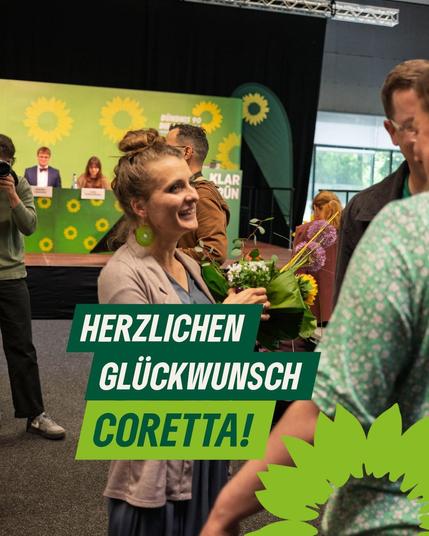 Bild zeigt die Halle eines grünen Parteitags - im Zentrum Coretta Storz - strahlend mit einem Blumenstrauß der Gratulation. Um Coretta stehen andere Parteimitglieder.