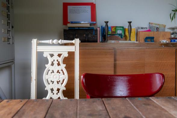 Photograph of a community cafe interior with a close-up of two chairs at a wooden table. One chair back is traditional, ornate and light coloured and beside it is a modern curved laminated wood chair back painted cherry red. The traditional table is made of wooden planks nailed to a frame. In the background against the wall is a modernist era wood sideboard, on top of which are various books, a music player, candlesticks, plants and office implements. A sign saying OPEN SOURCE CAFÉ is perched on top of a pile of books. To the left just visible is a metal cabinet. DeWAR social centre for the arts and citizen science, Amersfoort, Netherlands, 20 May 2024. Photo Cindy Kohtala
