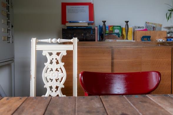 Photograph of a community cafe interior with a close-up of two chairs at a wooden table. One chair back is traditional, ornate and light coloured and beside it is a modern curved laminated wood chair back painted cherry red. The traditional table is made of wooden planks nailed to a frame. In the background against the wall is a modernist era wood sideboard, on top of which are various books, a music player, candlesticks, plants and office implements. A sign saying OPEN SOURCE CAFÉ is perched on top of a pile of books. To the left just visible is a metal cabinet. DeWAR social centre for the arts and citizen science, Amersfoort, Netherlands, 20 May 2024. Photo Cindy Kohtala