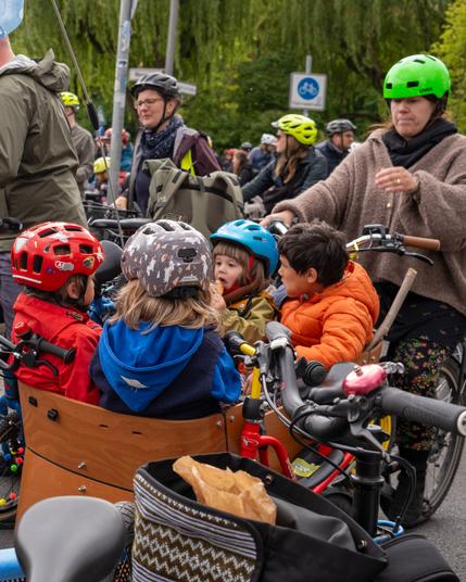 Kinder mit bunten Helmen sitzen in einem Lastenfahrrad, umgeben von Erwachsenen mit Fahrrädern