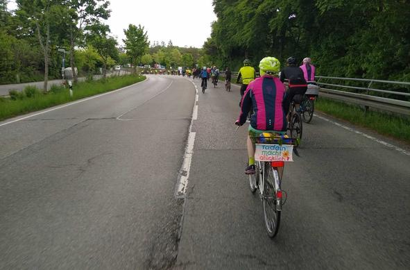 Fahrradtross in einer langen Kurve auf der Bundesstraße kurz vor Baierbrunn, vorne im Bild eine alten Dame auf dem Fahrrad, am Gepäckträger das Schild "Radeln macht glücklich."