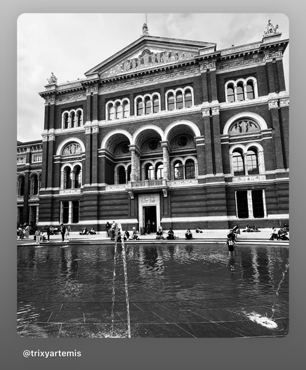 A black and white photograph of the courtyard at the Victoria and Albert Museum. The image captures an ornate red brick building with intricate architectural details, including arched windows and decorative stonework. In the foreground, a fountain sits in the middle of the open space, surrounded by a paved courtyard. The contrast between the dark tones of the brick and the lighter shades of the sky and stone features highlights the building’s historical elegance.