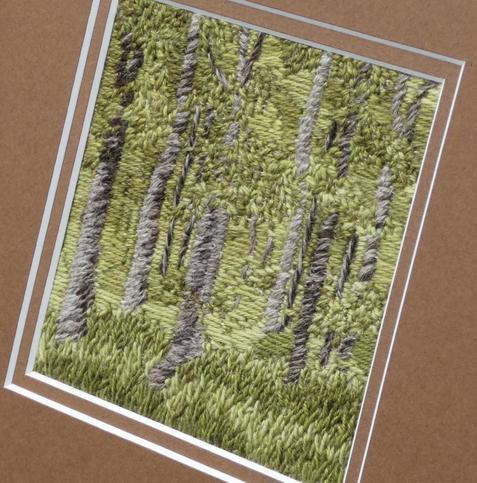 Stitched artwork depicting a summer woodland with light and shade. A very breen pallette with tall thin tree trunks in brown/grey. Close mounted in a brown double mount. Viewed on an angke to show texture.
