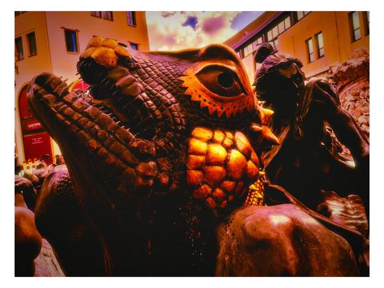 Das Bild zeigt eine detaillierte Bronzestatue, die einen alligatorähnlichen Krokodil mit einem auffälligen, großen Auge und einem aufgerissenen Maul darstellt. Die Skulptur ist in warmen, orangen-rote Tönen gehalten, die die Textur der Haut und die Konturen des Krokodils hervorheben. Im Hintergrund ist eine weitere Bronzestatue zu sehen, die einen menschlichen Figurenkörper mit einem Kopf zeigt, der möglicherweise eine traditionelle Kopfbedeckung trägt. Die Umgebung ist urban, mit Gebäuden im Hintergrund, die auf eine städtische Umgebung hinweisen. Der Himmel ist mit Wolken bedeckt, was dem Bild eine dramatische Atmosphäre verleiht. Die Statuen sind Teil einer öffentlichen Skulpturgruppe, die wahrscheinlich in einem öffentlichen Raum oder einem Park platziert ist.