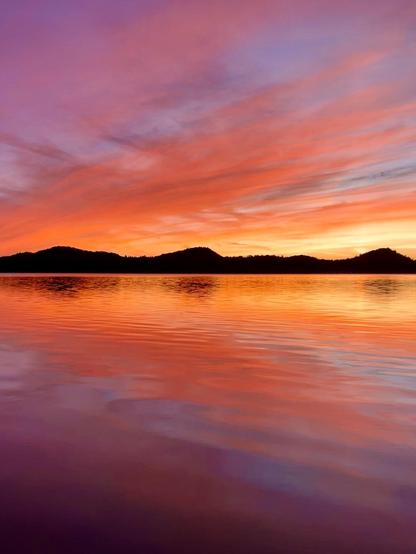 A serene sunset scene over a body of water, featuring vibrant orange, pink, and purple hues in the sky, reflecting softly on the water's surface. Silhouetted hills are visible in the background.