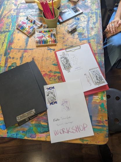 Auf einem bunt beschmierten Zeichentisch liegen zwei weiße DIN A4 -Zettel. Mit bunten Klebestreifen sind insgesamt vier Porträtfotos von Frauen aufgeklebt. Daneben sieht man die aufs Papier übertragenen Zeichnungen der Porträts. Auf dem Tisch liegen Stifte zum ausmalen der Bilder bereit. Auf einem der Zettel steht noch "Workshop"