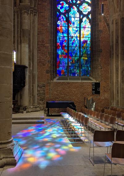 Im Seitenschiff einer Kirche schreibt die Sonne durch ein buntes Kirchenfenster und erzeugt ein buntes Farbenspiel auf dem gepflasterten Boden