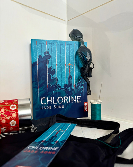 Livre posé contre du carrelage de douche, avec des lunettes de piscine, un maillot de piscine mais aussi une boite de thé ouverte, du fil bleu et une aiguille à coudre. Au mur, une ombre de queue de sirène. La couverture de Chlorine montre une sirène dans une piscine de natation dont la queue, ensanglantée, dégage un nuage d’encre derrière elle.