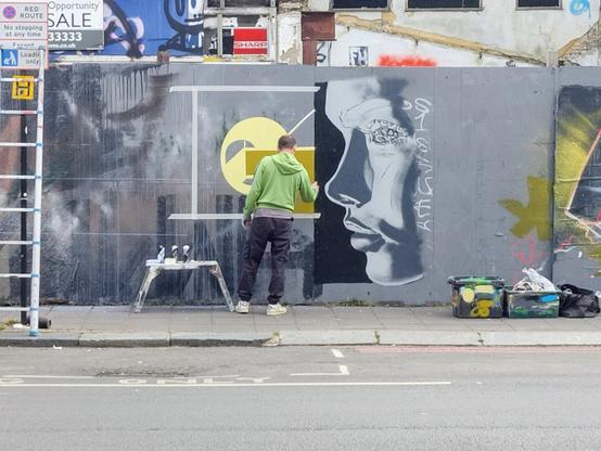 Man in green hoodie spray painting urban mural of abstract shapes yellow and green and a grey woman's face from the side on a grey background.