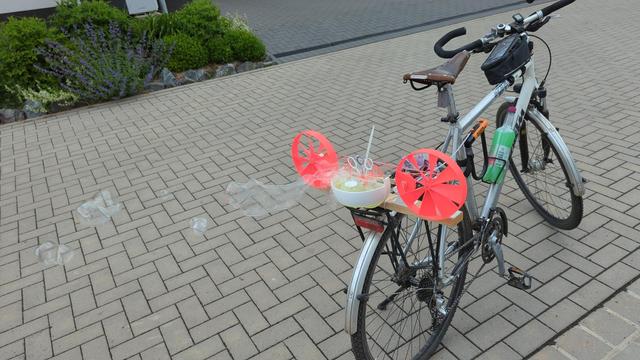 Eine Seifenblasenmaschine ist auf dem Gepäckträger vom Fahrrad befestigt. Der Wind treibt über 2 rote Windräder die Machine an.