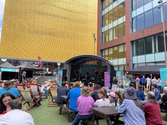 An outdoor event featuring a live band on stage, with an audience seated on deck chairs and at tables. Colorful banners are present, and a distinctive yellow building serves as a backdrop. Various vendors and food stands are visible in the background.