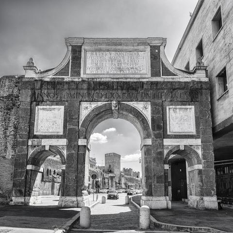 Black and white processing of a photo of the Arch of Sixtus V in Rome Italy. Though the arch can be seen a modern street with cars but bordered with ancient Roman walls.