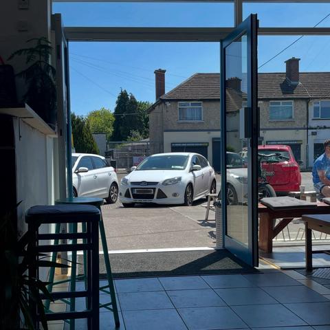 A view through an open door of a cafe seating area, showing a sunny outdoor scene with parked cars. Green trees and buildings are visible in the background, along with a person seated at a table outside.