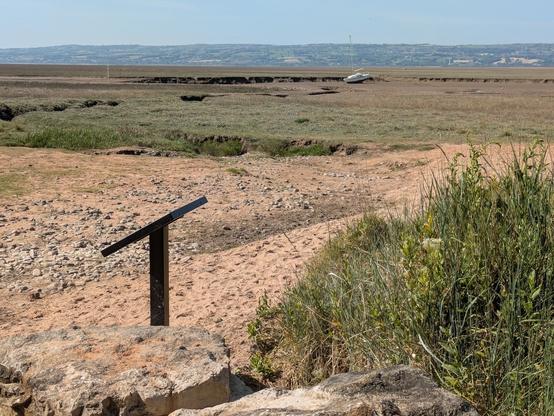 A barren landscape featuring a dry, grassy terrain with a distant beached yacht and a small explainer in the foreground and Wales beyond.
