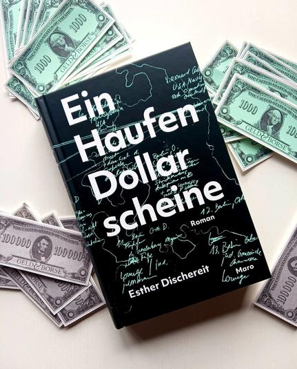 Foto des Buches, auf dessen schwarzem Cover mit weißer Schrift in quasi unleserlicher helltürkisfarbener Handschrift wohl Orts- und Personennamen stehen sowie etwas wie eine grob umrissene Karte zu sehen ist. Das Buch liegt auf einer weißen Fläche, daneben liegen türkisfarbene und graue Spielgeldscheine aus einem Spiel namens »Geld & Börse« im Wert von 1.000 und 100.000 Einheiten, auf den türkisfarbenen ist George Washington, auf den grauen Abraham Lincoln zu sehen.