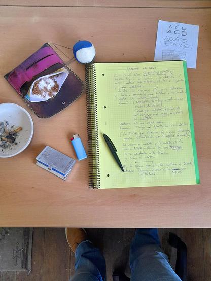 Sobre una mesa, una libreta de hojas amarillas con un escrito que espero que no se pueda leer. Una petaca de tabaco de liar, una lata de papel de fumar, un mechero y cenicero con restos de uso (colillas). Una pequeña pelota para apretar con la mano, de tela cosida en forma de yin-yan y una hoja de papel suelta que prefiero que no se lea, por curro.