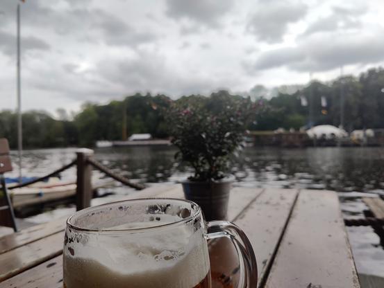Foto quer über einen Tisch im einem Biergarten am Wasser, das Bierglas ist im Fokus, jedoch ist nur die Schaumkrone zu sehen. Im Hintergrund unscharf sieht man Wasser, Hausboote und Wald. Der Regen ist kaum zu erkennen.