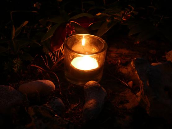 Das Foto zeigt ein Glas mit einem Teelicht zwischen Steinen. Das Foto ist bei Dunkelheit aufgenommen, daher verschwindet die Umgebung in der Dunkelheit. Mann kann im Glas gut den gespiegelten Docht und die Flamme erkennen.