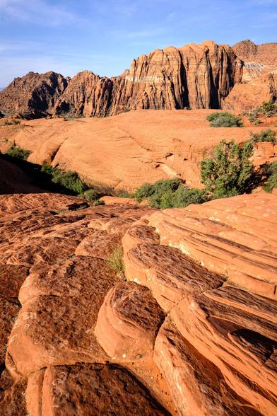 Layered red sandstone formations with ridges and grooves, sparse vegetation, and distant rugged cliffs under a blue sky.
