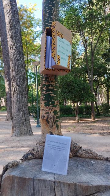 Hay un buzón hecho con una caja de cartón y decorado con pintas de leopardo, tiene un cartel escrito a mano: coge uno y vuelve a cerrar el buzón. Está atado a un tronco fino de un árbol, que es un brote de un tronco anteriormente talado. En el trozo más abajo del tronco talado hay un minizine de pie que pone ¿Y ahora qué hacemos? Al fondo se ven más troncos y árboles, es un parque y el suelo es de tierra