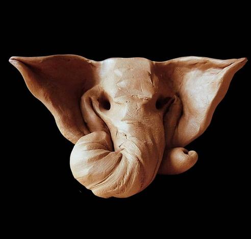 This image shows a clay or terracotta sculpture of an elephant head against a black background. The sculpture captures just the head, featuring large, distinctive ears that spread outward, a trunk that curves slightly to one side, and small tusks. The piece has a natural earthy brown color with subtle variations in tone. The sculpture appears to be handcrafted with visible textures and details that give it character, including subtle indentations and creases in the clay. The style is somewhat stylized while maintaining recognizable elephant features, creating an artistic representation rather than a strictly realistic one.